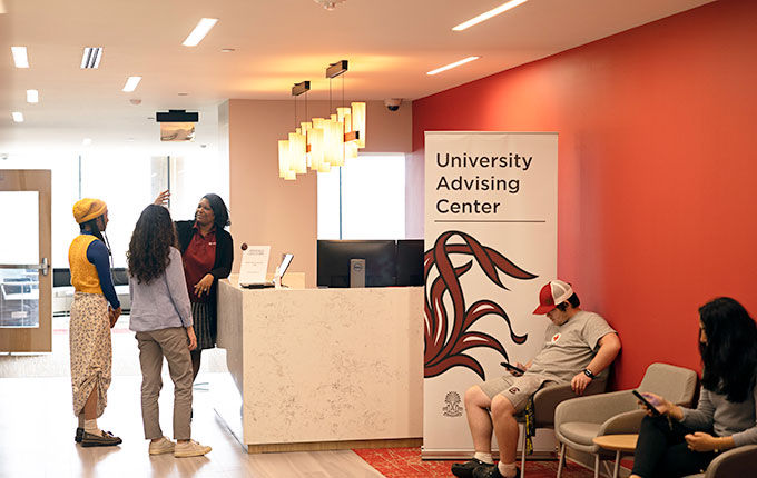 An African American academic advisor stands at a check in desk in the University Advising Center talking with 2 students