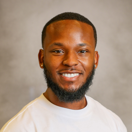 Professional headshot of a smiling person with closely cropped black hair and a neatly trimmed beard wearing a light gray crew-neck shirt. The person is facing the camera and is wearing small stud earrings. The photo is set against a soft gray studio background with warm, even lighting, creating a friendly, approachable, and professional appearance.
