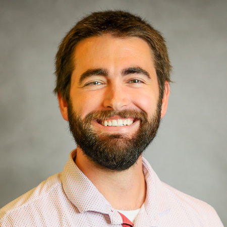 Professional headshot of a smiling person with short dark brown hair and a full beard, wearing a light-colored button-up shirt with a subtle pattern over a red undershirt. They are facing the camera directly with a broad, friendly smile. The portrait is set against a softly blurred gray background, creating a warm, approachable, and professional appearance.