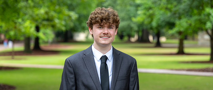 A graduating senior in a suit stands outdoors on a green campus lawn.