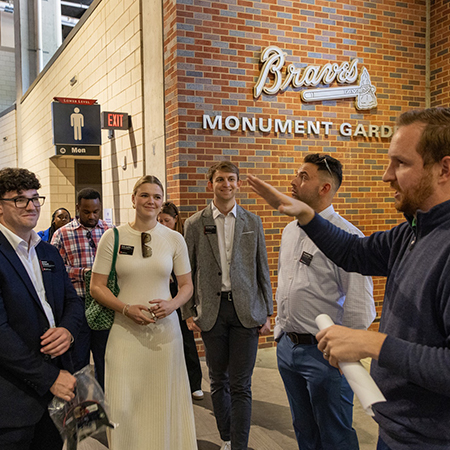 A tour guide gestures while speaking to a group of professionally dressed students standing near the Braves Monument Garden inside Truist Park.