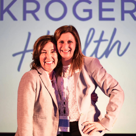 Two women pose for photo in front of Kroger Health sign