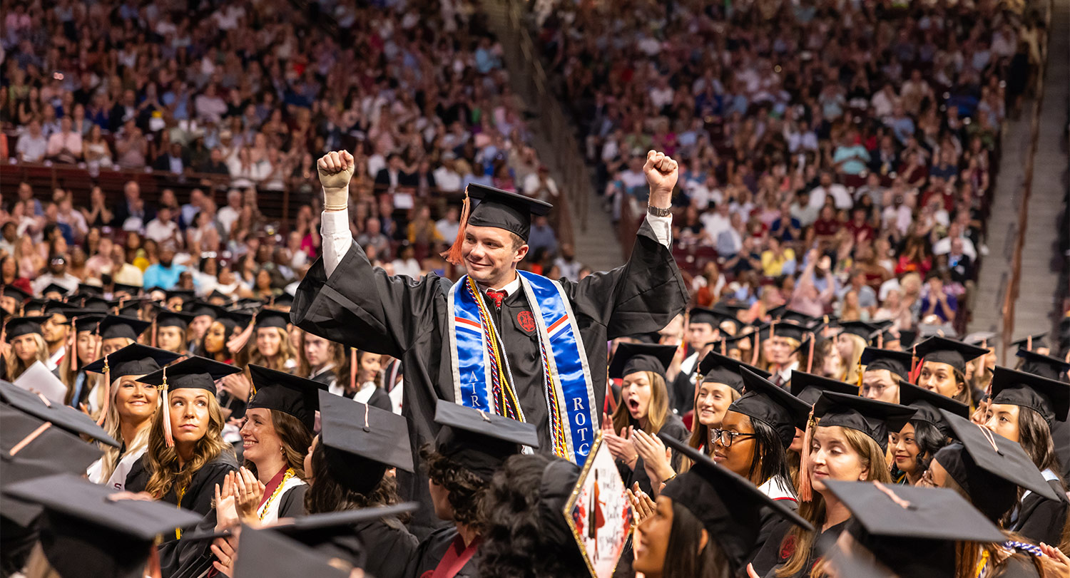 Student in cap and gown stands with fists in the air at commencement ceremony