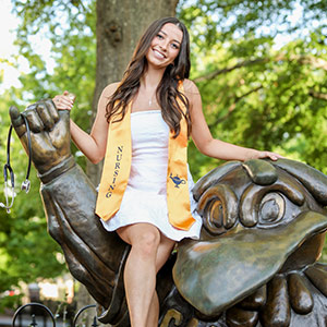 Student poses on Cocky statue in graduation sash